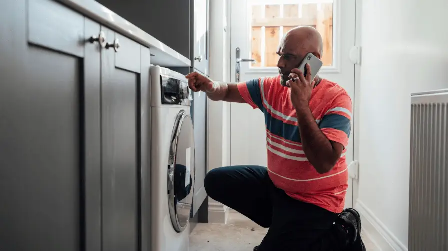 Man kneeling in front of his washer machine, trying to get it to work, while speaking on his phone.